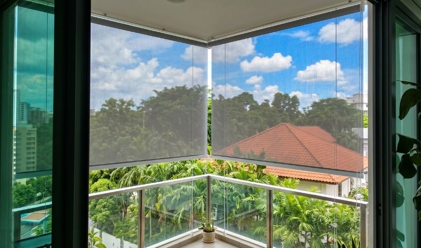Grey mesh outdoor roller blinds installed on a corner Singapore balcony with glass balustrade and tropical garden view